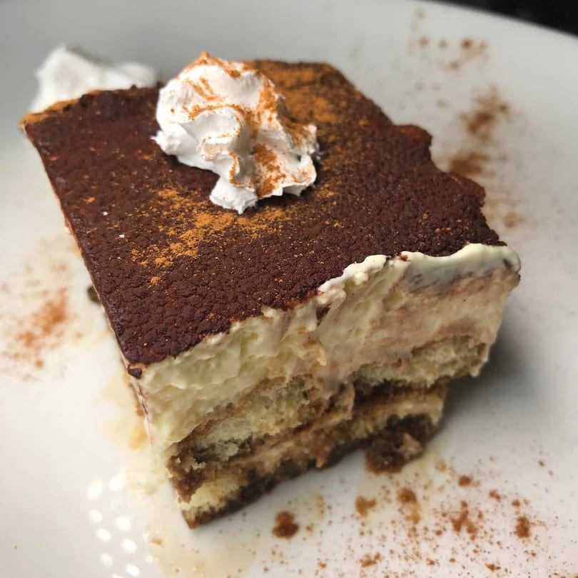 A slice of tiramisu with whipped cream and cinnamon dusting on a white plate.