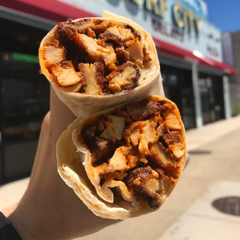 Hand holding a chicken shawarma wrap, cut in half, with the Surf City Italian Restaurant in the background.